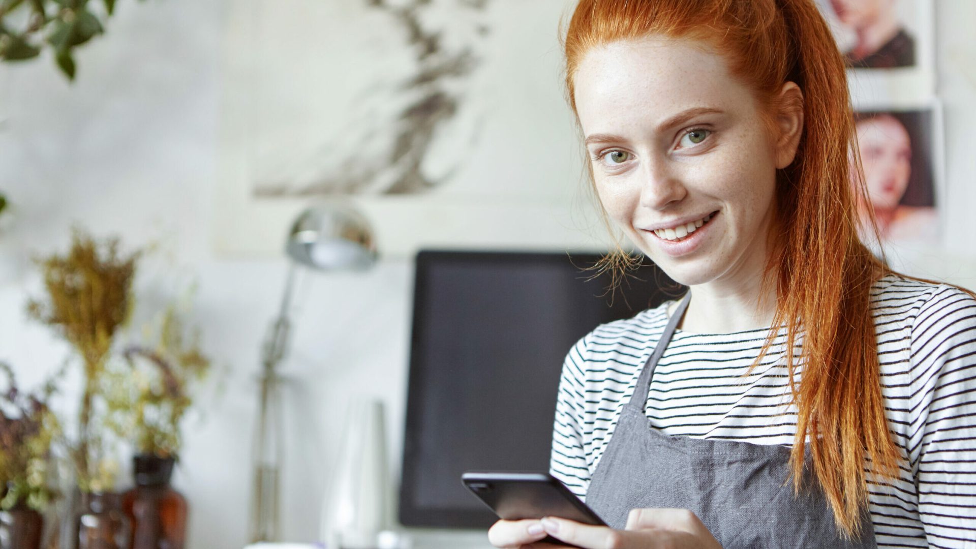 Close up portrait of cheerful cute young woman of creative occupation wearing her long ginger hair in ponytail typing text message using her mobile phone, looking and smiling broadly at camera